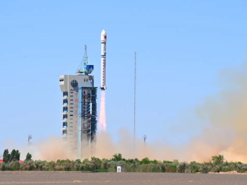 A white Long March 2D rocket lifts off from the Jiuquan Satellite Launch Center. The rocket ascends into a clear blue sky with exhaust plumes trailing below, as the launch tower and desert landscape appear in the foreground.