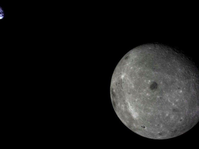 Image of the moon's far side with Earth in the distant background, taken from lunar orbit. The lunar surface appears gray and cratered, while Earth is shown as a small blue-and-white sphere against the blackness of space.