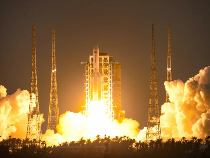 A dramatic nighttime launch of China's Long March 5B rocket from the Wenchang Space Launch Center on December 16, 2024. Bright orange flames and thick clouds of white smoke illuminate the launchpad as the rocket ascends into the sky, carrying the first 10 satellites for the Guowang ('national net') low Earth orbit communications megaconstellation. Tall support towers frame the scene against the dark backdrop, highlighting the power and scale of the mission.