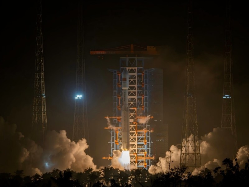 A Long March 8 rocket lifts off from Hainan Commercial Launch Site at night, illuminating the surroundings with bright flames and thick clouds of smoke. The launch pad structure and lightning towers frame the ascending vehicle against the dark sky.