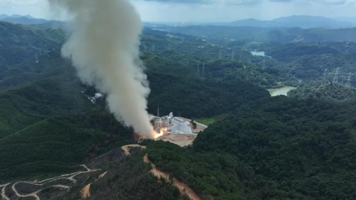 Aerial view of CAS Space's Kinetica-2 first-stage hot fire test, showing flames and thick smoke rising from a test stand nestled in forested hills.