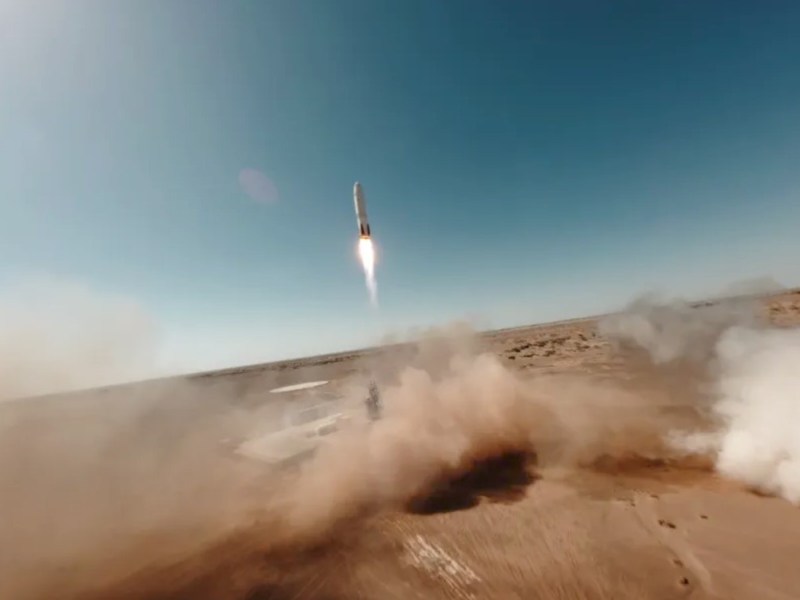 Sand blows off a desert landing pad as a Nebula-1 first stage makes a powered descent from clear blue skies towards a landing pad.