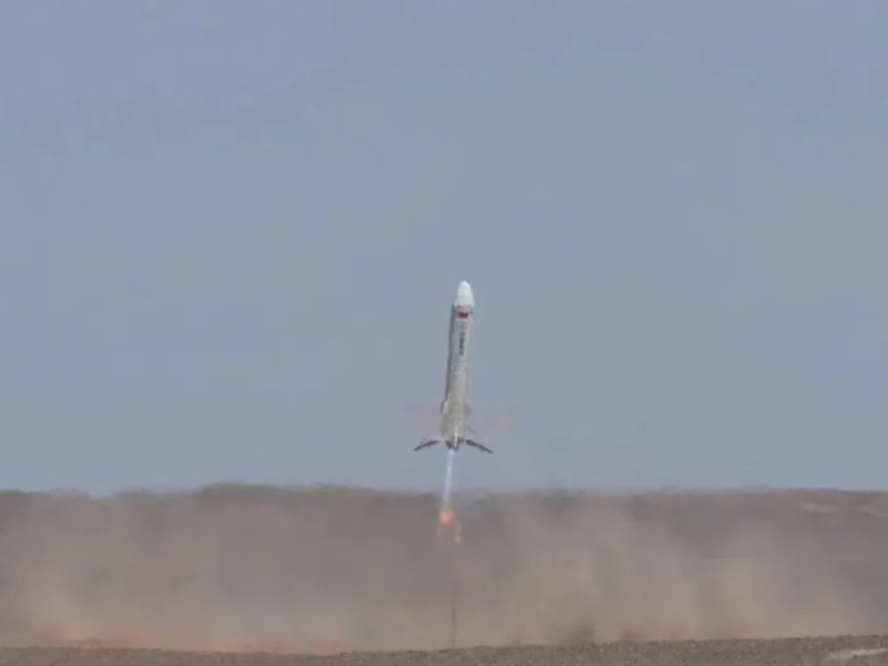 A SAST (Shanghai Academy of Spaceflight Technology) vertical takeoff, vertical landing (VTVL) rocket prototype during a test flight, kicking up dust from the ground. The rocket's engines emit a bright flame, indicating an active and controlled descent. The test was conducted in a desert-like environment under clear skies.