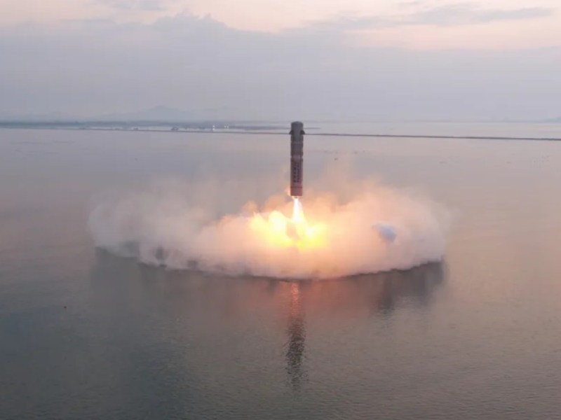 A tall, cylindrical rocket is captured in the final moments of a vertical descent, hovering just above the surface of calm coastal waters during sunrise. Bright orange engine flames and thick plumes of white smoke billow outward as the rocket slows its descent for a soft splashdown. The scene is framed by a hazy sky, with distant land barely visible on the horizon. The water below reflects the rocket's light, and the atmosphere appears serene despite the powerful landing burn.