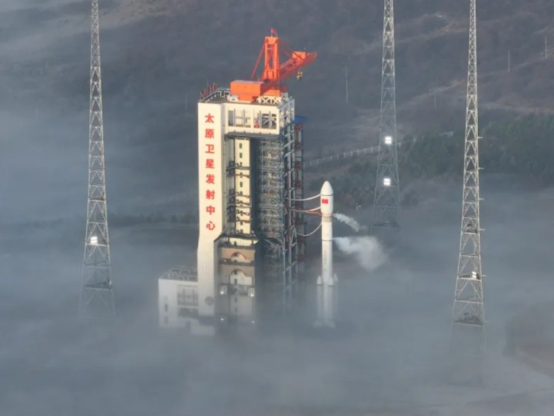 A Long March 6A rocket stands on the launch pad at China's Taiyuan Satellite Launch Center, surrounded by dense morning fog and lightning towers. Venting is visible from the rocket as it prepares for liftoff, with the launch tower displaying Chinese characters reading "Taiyuan Satellite Launch Center."