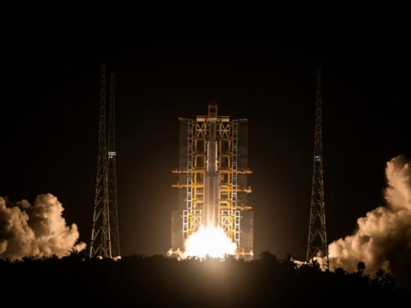 A Long March 7A rocket lifts off at night from China's Wenchang Satellite Launch Center, illuminating the launch pad with bright exhaust flames and casting shadows on surrounding support structures and trees.