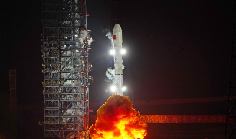 A Long March 3B rocket launches ChinaSat-9C from Xichang Satellite Launch Center at night, with flames and smoke billowing from the base and the illuminated rocket lifting off against a dark sky.